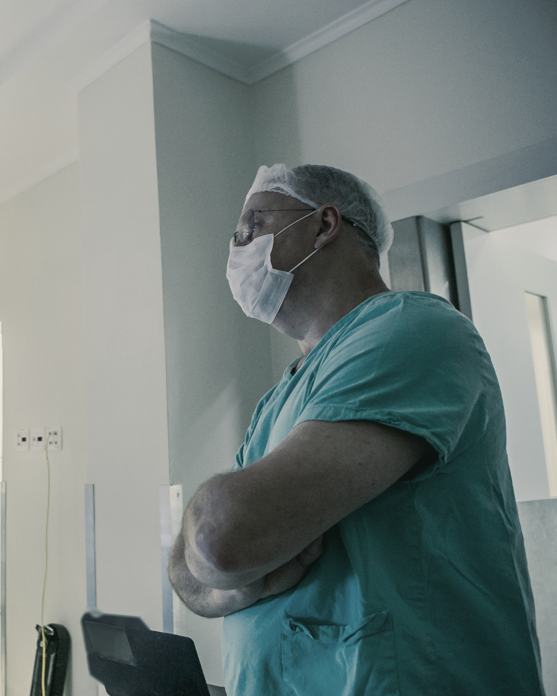Dr. Fábio Luiz Waechter no bloco cirúrgico, de touca, máscara e roupa de bloco, braços cruzados, acompanhando a cirurgia no monitor